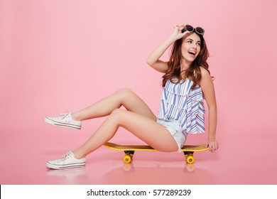 Happy attractive young woman in sunglasses sitting on skateboard over pink background - Shutterstock ID 577289239