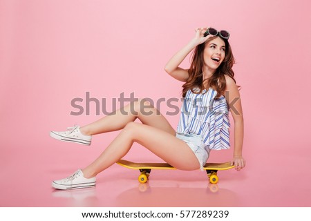 Happy attractive young woman in sunglasses sitting on skateboard over pink background