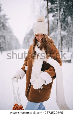 Happy attractive young woman in warm woolen jacket, white scarf, hat and mittens posing with crochet net bag of tangerines, walking in snowy winter forest