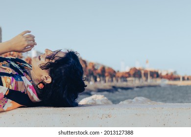 Happy Authentic Brunette Curly Hair Woman Laying Down On The Coast Side And Laughing With Sea And City In The Background