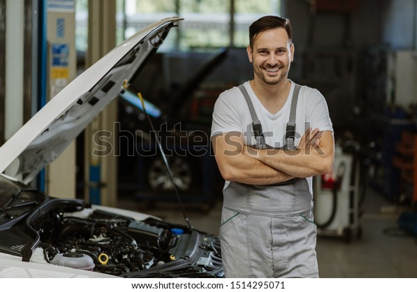 Happy auto mechanic standing at\
workshop with his arms crossed and looking at\
camera
