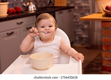 A Happy Baby 10-12 Months Old Eats Milk Porridge With A Spoon. Portrait Of A Happy Girl In A Highchair In The Kitchen