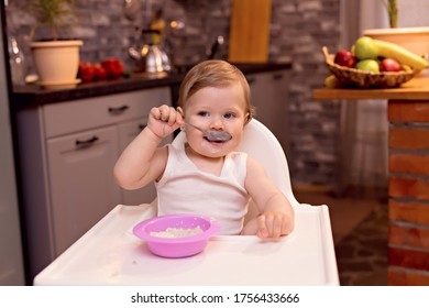 A Happy Baby 10-12 Months Old Eats Milk Porridge With A Spoon. Portrait Of A Happy Girl In A Highchair In The Kitchen