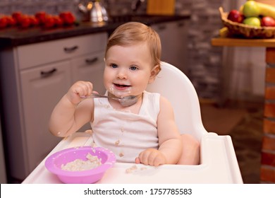 A Happy Baby 10-12 Months Old Eats Milk Porridge With A Spoon. Portrait Of A Happy Girl In A Highchair In The Kitchen