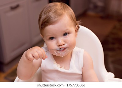 A Happy Baby 10-12 Months Old Eats Milk Porridge With A Spoon. Portrait Of A Happy Girl In A Highchair In The Kitchen