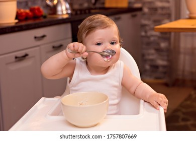 A Happy Baby 10-12 Months Old Eats Milk Porridge With A Spoon. Portrait Of A Happy Girl In A Highchair In The Kitchen