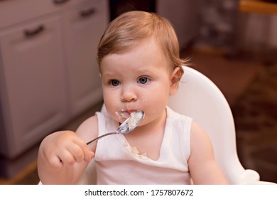 A Happy Baby 10-12 Months Old Eats Milk Porridge With A Spoon. Portrait Of A Happy Girl In A Highchair In The Kitchen