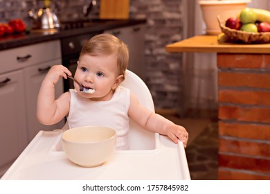 A Happy Baby 10-12 Months Old Eats Milk Porridge With A Spoon. Portrait Of A Happy Girl In A Highchair In The Kitchen