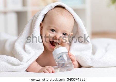 Happy baby boy drinks water from bottle wrapped towel after bath