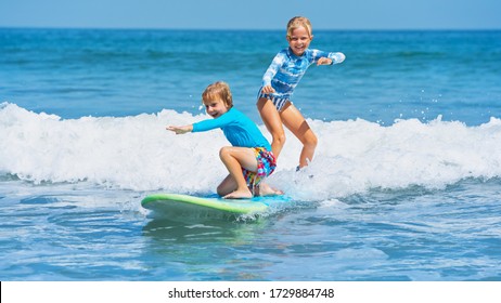 Happy Baby Boy And Girl - Young Surfers Ride With Fun On One Surfboard. Active Family Lifestyle, Kids Outdoor Water Sport Lessons, Swimming Activity In Surf Camp. Sea Beach Summer Holiday With Child.