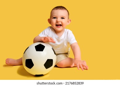 Happy Baby Boy Plays With A Soccer Ball On A Yellow Studio Background, Copy Space. A Smiling Child Learns To Play Football With A Big Ball. Kid Age One Year