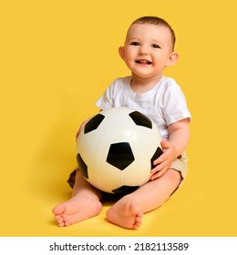 Happy Baby Boy Plays With A Soccer Ball On A Yellow Studio Background, Copy Space. A Smiling Child Learns To Play Football With A Big Ball. Kid Age One Year