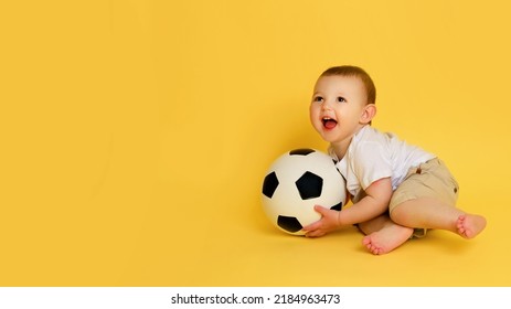 Happy Baby Boy Plays With A Soccer Ball On A Yellow Studio Background, Copy Space. A Smiling Child Learns To Play Football With A Big Ball. Kid Age One Year