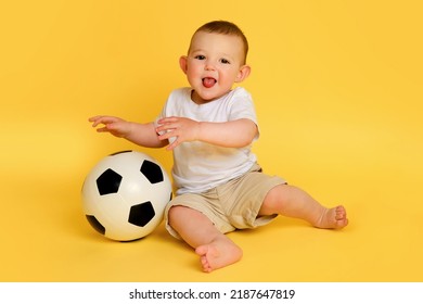 Happy Baby Boy Plays With A Soccer Ball On A Yellow Studio Background, Copy Space. A Smiling Child Learns To Play Football With A Big Ball. Kid Age One Year