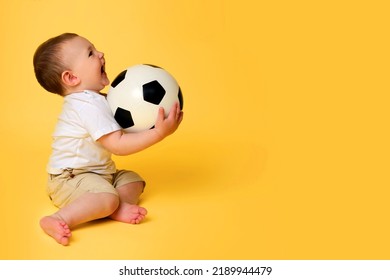 Happy Baby Boy Plays With A Soccer Ball On A Yellow Studio Background, Copy Space. A Smiling Child Learns To Play Football With A Big Ball. Kid Age One Year