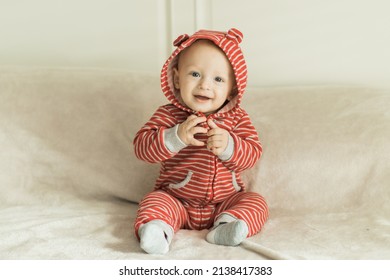 Happy Baby Boy Smiling While Sitting On A Beige Blanket With A White Headboard. He Is Smiling And Wearing A Cute One Piece Outfit Red Stripes With Little Puppy Ears Hoodie And Gray Socks.