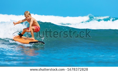 Happy baby boy - young surfer learn to ride on surfboard with fun on sea waves. Active family lifestyle, kids outdoor water sport lessons, swimming activity in surf camp. Summer vacation with child.