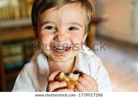 Happy baby eats sweet chocolate cake. He wears a white summer shirt and is sitting in his highchair to eat. Children's concept