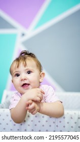 Happy Baby Girl With Brown Eyes Looking Away With Smile While Standing In Crib Against Colorful Ornamental Wall In Bedroom