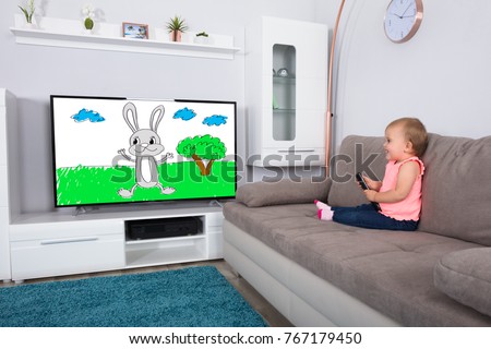Happy Baby Girl Sitting On Sofa Watching Cartoon On Television