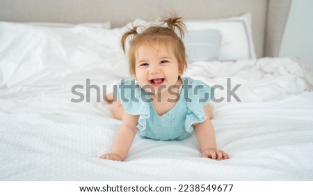Happy baby girl smiling in blue suit pajama in white bedroom lying on bed looking at camera. Cute child with pigtails. Childhood, babyhood, people concept. Copy space fro advertisement