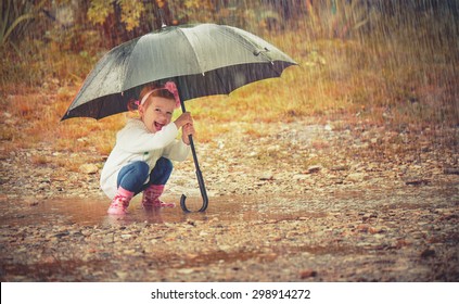 Happy Baby Girl With An Umbrella In The Rain Runs Through The Puddles Playing On Nature