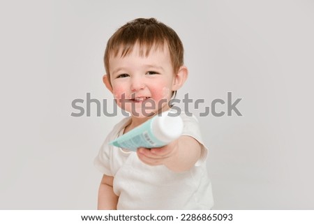 Happy baby holding a tube of cream on a studio white background. Smiling child plays with a package of ointment. Kid about two years old (one year nine months)