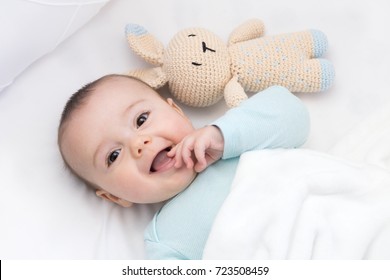 Happy Baby Newly Awake In His Crib And With His Bunny Doll. Light Blue Pajama And White Bed Sheets.