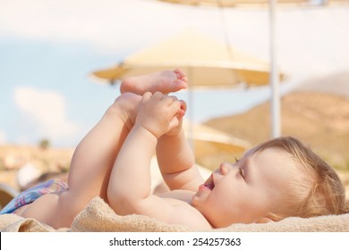 Happy Baby On The Beach Sunbed. 8 Month Old Kid Lying On A Sun Lounger And Playing With Her Feet. Summer Holidays Concept.