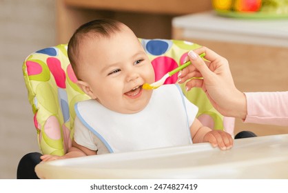 A happy baby sits in a colorful high chair while being fed with a spoon by an adult. The scene takes place in a bright home kitchen, suggesting a casual and joyful mealtime. - Powered by Shutterstock