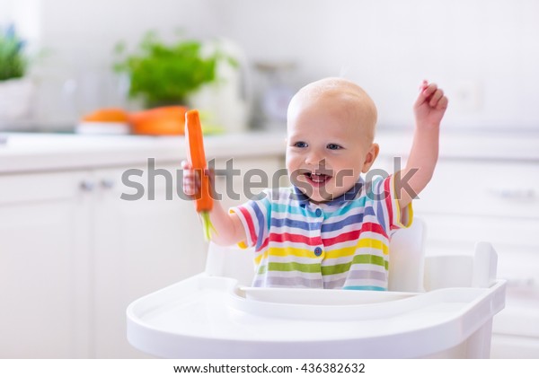 Happy baby sitting in high chair eating carrot\
in a white kitchen. Healthy nutrition for kids. Bio carrot as first\
solid food for infant. Children eat vegetables. Little boy biting\
raw vegetable.