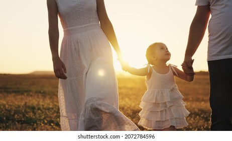 Happy Baby Smiling At Sunset With Parents, Mother, Father And Child Walk In The Sun, Hold Kid By Hands, Young Family, Taking Care Of Their Little Daughter, Family Walk In Evening Park In Orange Light