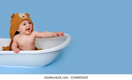 Happy baby toddler boy with hood bathrobe shows his hand in a white tub on a studio blue background. A smiling child at the age of one year, copy space