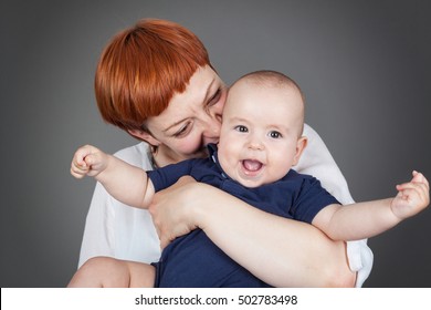 Happy Baby-Close Up Of Mother Holding And Kissing A Joyfull Baby Boy Who Is Laughing With Both Arms Stretched Out, Isolated On Grey With Vignette, Horizontal