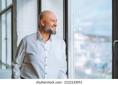 Happy Bald Bearded Mature Businessman In White Shirt Standing Near Panoramic Window And Enjoying City View. Successful Smiling Middle-aged Male CEO Executive In Office, Selective Focus