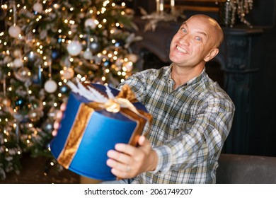 Happy Bald Man In A Shirt Holding A Blue Box With A Gift On The Background Of A Christmas Tree