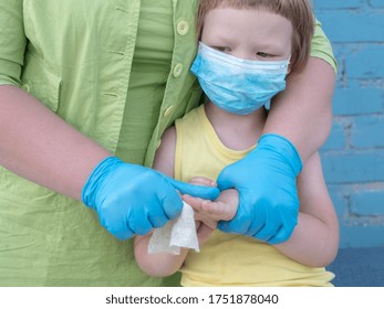 Happy Bald Mother In Green Dress, Medical Mask And Blue Gloves Holds Bottle With Disinfectant In Her Hands. Woman Teaches Her Child To Use Sanitizer For Reliable Protection Against Viruses