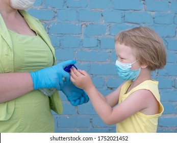 Happy Bald Mother In Green Dress, Medical Mask And Blue Gloves Holds Bottle With Disinfectant In Her Hands. Woman Teaches Her Child To Use Sanitizer For Reliable Protection Against Viruses