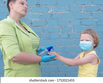 Happy Bald Mother In Green Dress, Medical Mask And Blue Gloves Holds Bottle With Disinfectant In Her Hands. Woman Teaches Her Child To Use Sanitizer For Reliable Protection Against Viruses