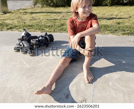 Happy barefoot boy taking a break from rollerblading sitting on the pavement on a sunny day. Active lifestyle. Happy childhood