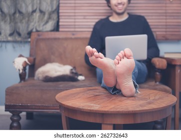 A happy barefoot man is resting his feet on a coffee table at home while working on his laptop, there is a cat sleeping next to him