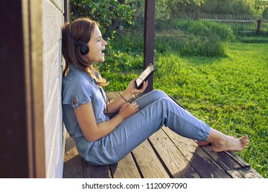 Happy Barefoot Teen Girl Wearing Headphones Listening To Music From A Smartphone And Singing Outdoors In Warm Summer Evening Sitting On A Porch Of Country House                                