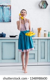 happy barefoot young woman talking on retro yellow telephone and laughing in kitchen  