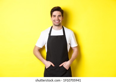 Happy barista in black apron looking at camera. Coffee shop owner wearing cafe uniform and smiling, standing over yellow background