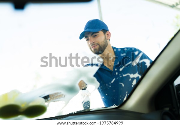 Happy\
to be doing a good job at the car wash. Handsome latin worker\
cleaning the car windshield with soap and a\
sponge