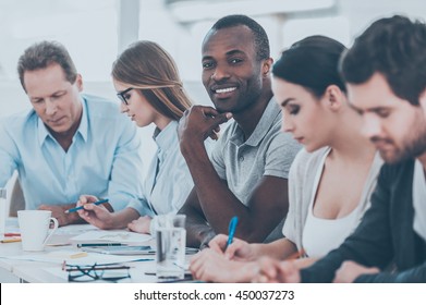 Happy to be a part of grat team. Group of business people sitting in a row at the table while handsome African man looking at camera and smiling - Shutterstock ID 450037273