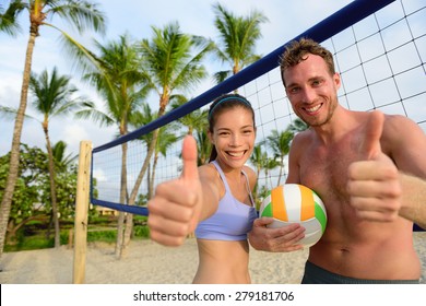 Happy beach volleyball players thumbs up. Excited smiling man and woman with beach volley ball giving thumbs up success hand sign looking at camera. Asian woman, Caucasian man.