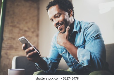 Happy bearded African businessman using phone while sitting on sofa at his modern home.Concept of young people working mobile devices.Blurred background