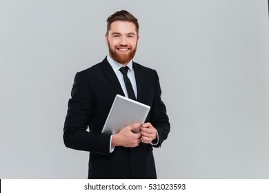 Happy Bearded Business Man In Black Suit Holding Tablet Computer And Looking At Camera In Studio. Isolated Gray Background