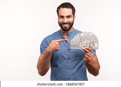Happy bearded businessman in blue shirt pointing finger at dollar cash in his hand, looking at camera with toothy smile, good salary, payments. Indoor studio shot isolated on white background